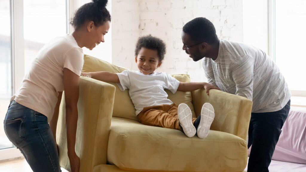 Happy family moving a yellow armchair, with a smiling child sitting on it as the parents lift it together in a bright, modern living room.